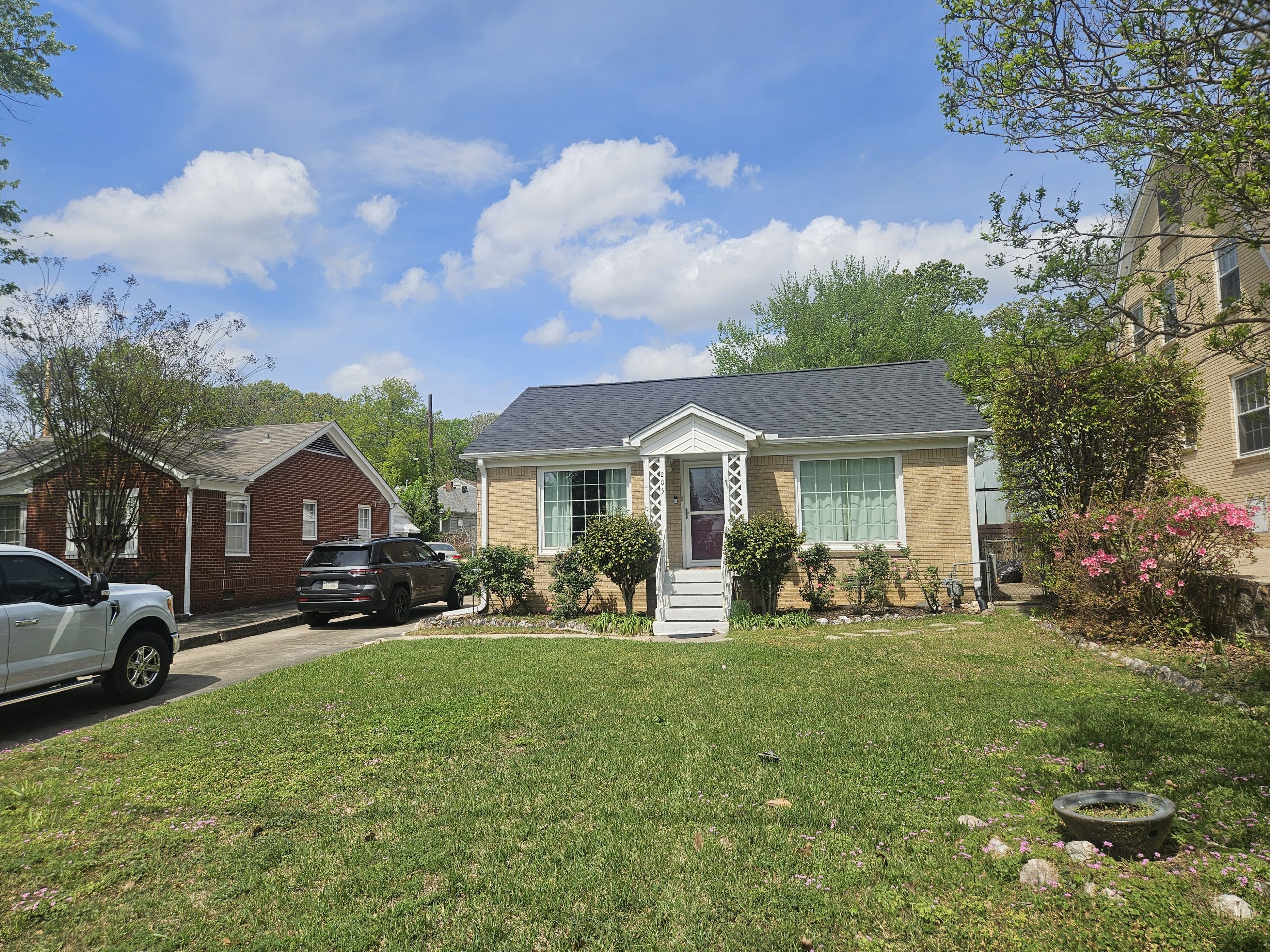 A front view of a single-story brick bungalow-style house with a dark grey shingle roof and white porch columns, located in Benton, Arkansas, on a sunny day.